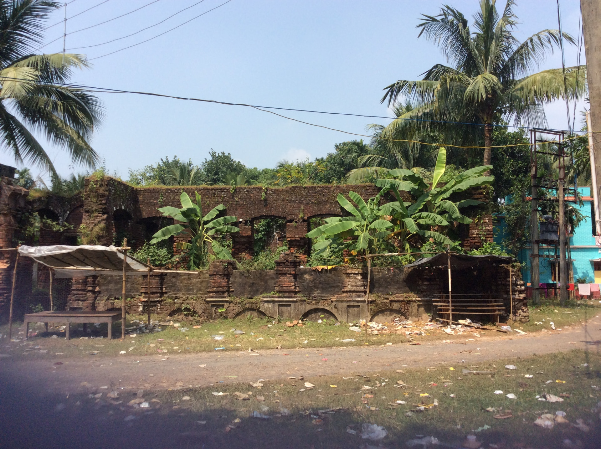 The roofless red-brick walls of an old house with a row of arched windows, banana plants growing inside, beside a village road