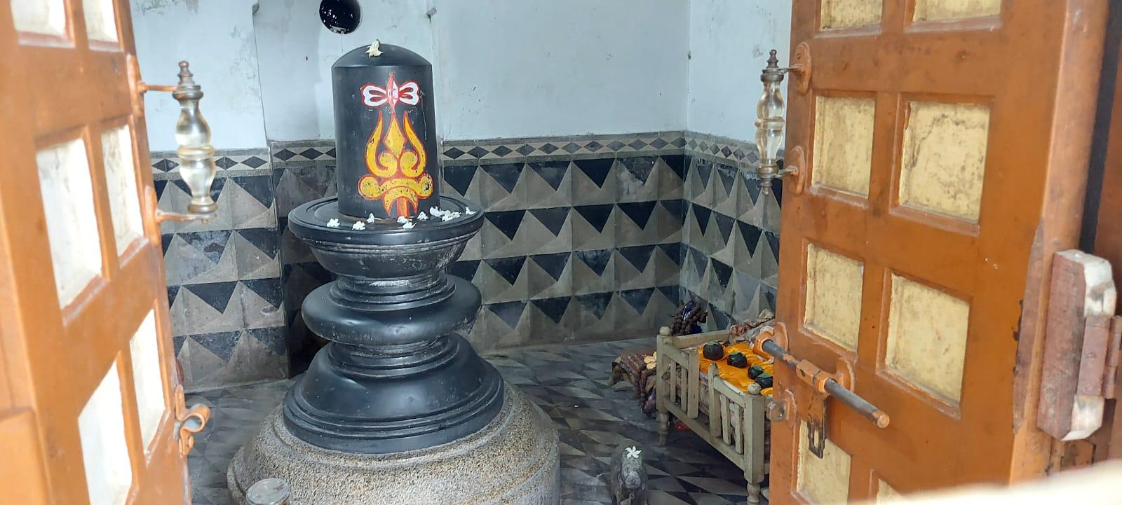 Buro Shiv: a large black stone Shiva lingam marked with a red and yellow tilak, flowers on its base, seen through the open wooden temple doors